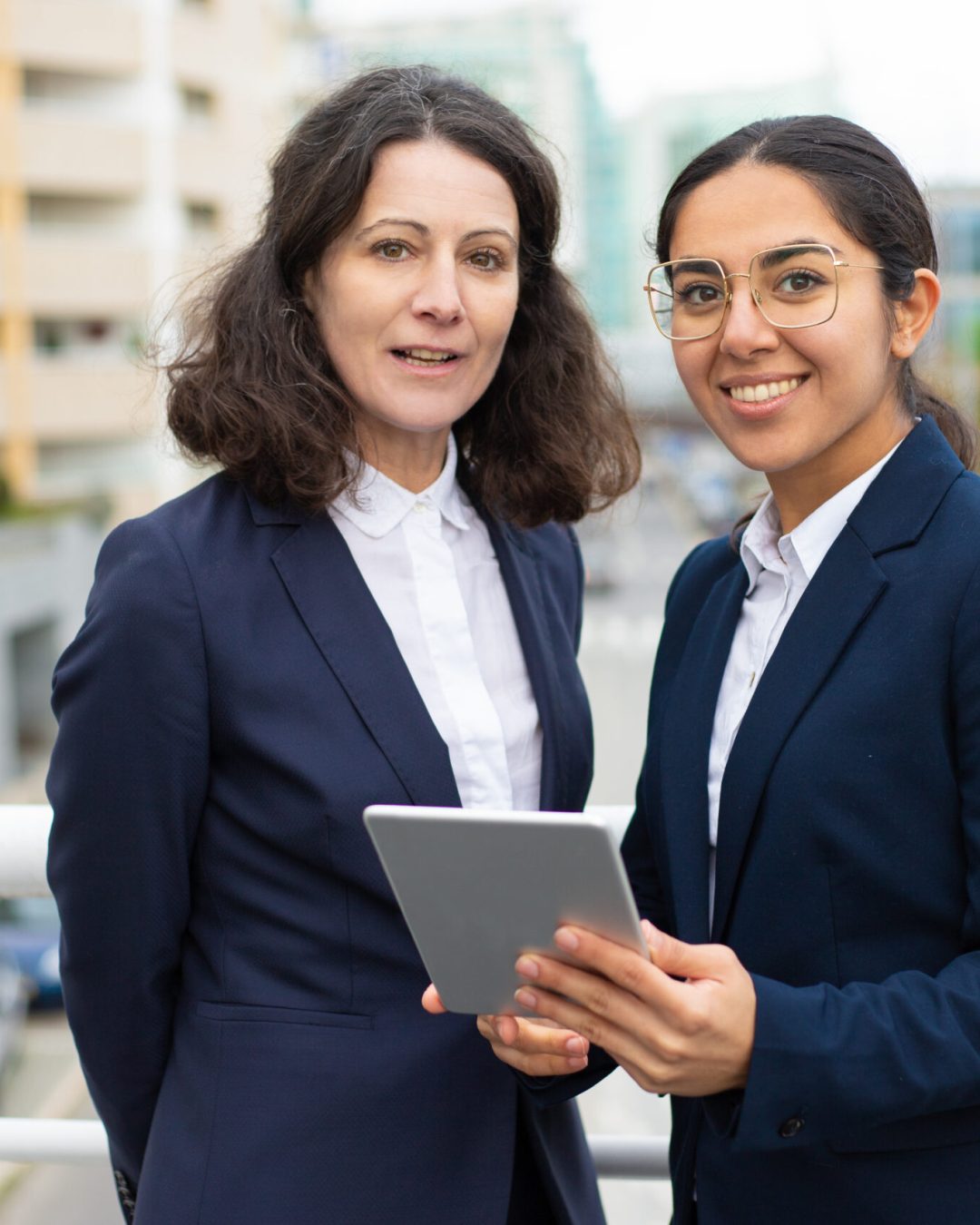 Content businesswomen with digital tablet. Cheerful professional female colleagues standing with digital tablet and smiling at camera at street. Wireless technology concept